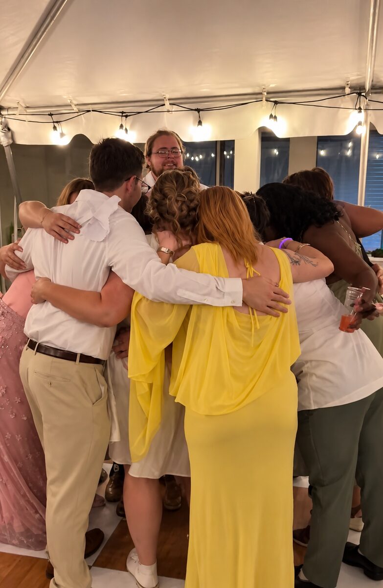 Ethan’s wedding party in a group hug under the tent lights